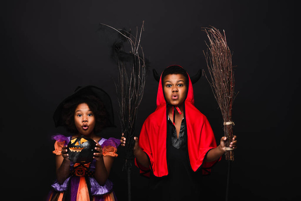 Afričanky americká dívka v halloween kostým držení vyřezávané dýně v blízkosti bratra s košťaty izolované na černé  - Fotografie, Obrázek