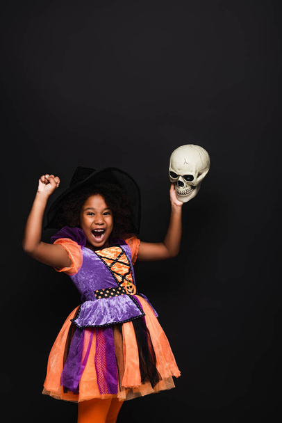 amazed african american kid in witch halloween costume holding skull isolated on black - Foto, Imagen
