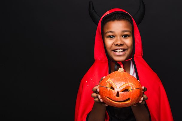 cheerful african american boy in devil halloween costume holding carved pumpkin isolated on black - Foto, imagen