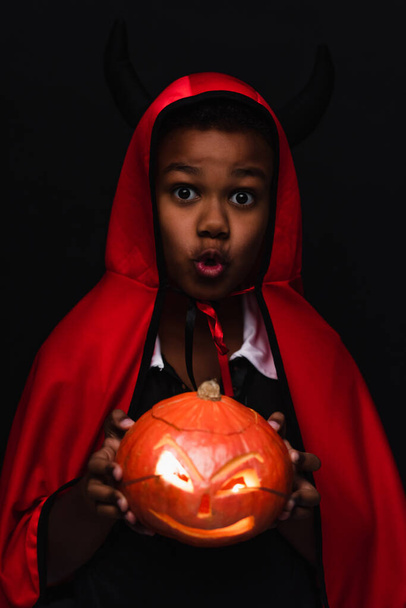 spooky african american boy in devil halloween costume holding carved pumpkin isolated on black - Fotó, kép
