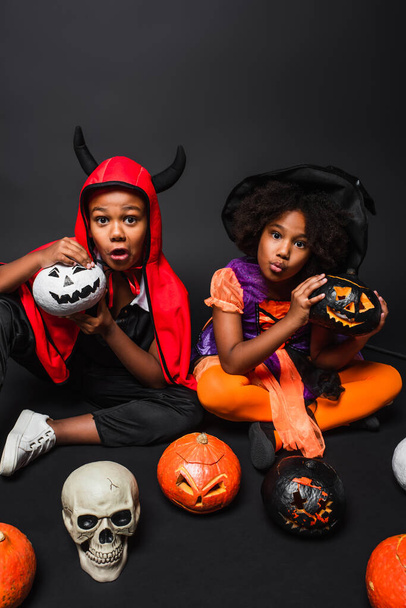 Gruselige afrikanisch-amerikanische Kinder in Halloween-Kostümen mit Totenkopf und Besen in der Nähe von Kürbissen auf schwarz  - Foto, Bild