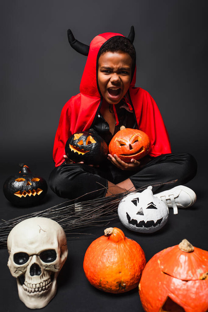 african american boy in halloween devil costume screaming while holding carved pumpkins near skull on black  - Foto, afbeelding