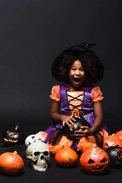 amazed african american girl in witch costume and pointed hat near carved pumpkins on black - Foto, Imagem