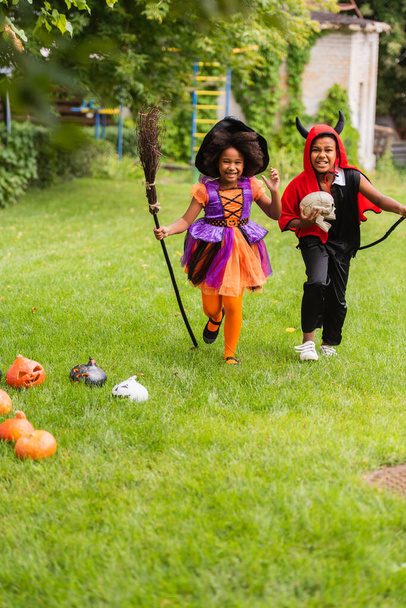 onnellinen afrikkalainen amerikkalainen lapset halloween puvut tilalla luudat ja käynnissä nurmikolla  - Valokuva, kuva