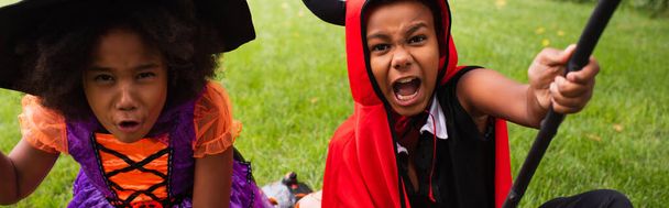 african american siblings in halloween costumes holding brooms and playing on backyard, banner - Foto, Bild