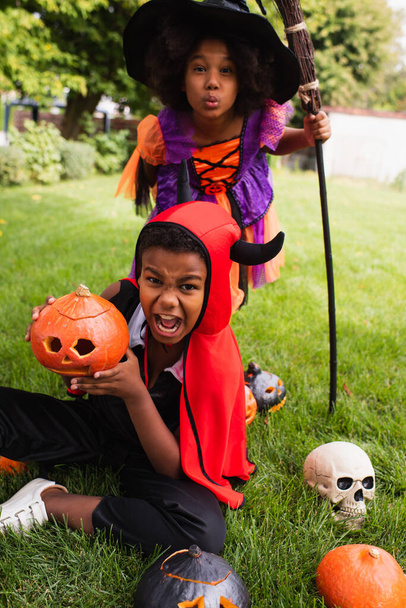 scary african american siblings in halloween costumes screaming near carved pumpkins on backyard - Foto, imagen