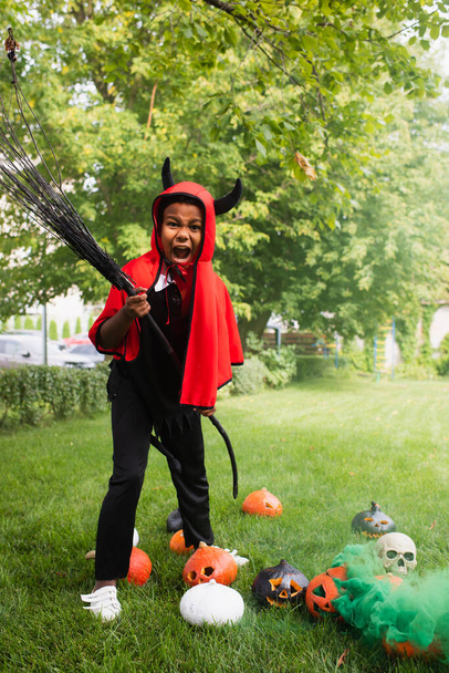 scary african american boy in devil halloween costume holding broom near pumpkins on lawn  - Foto, Imagem