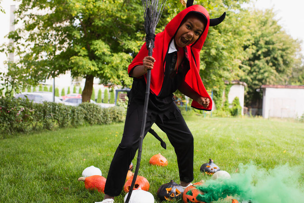 Gruseliges afrikanisch-amerikanisches Kind im Teufel-Halloween-Kostüm mit Besen neben Kürbissen auf dem Rasen  - Foto, Bild