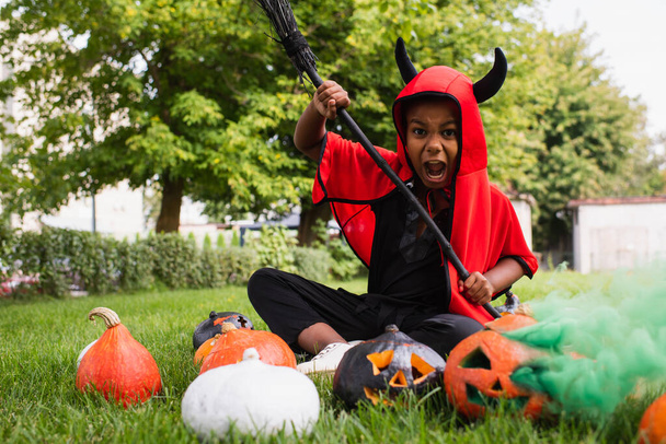 straszny afrykański amerykański dzieciak w diabelskim kostiumie halloween krzycząc trzymając miotłę w pobliżu dyń i siedząc na trawniku  - Zdjęcie, obraz