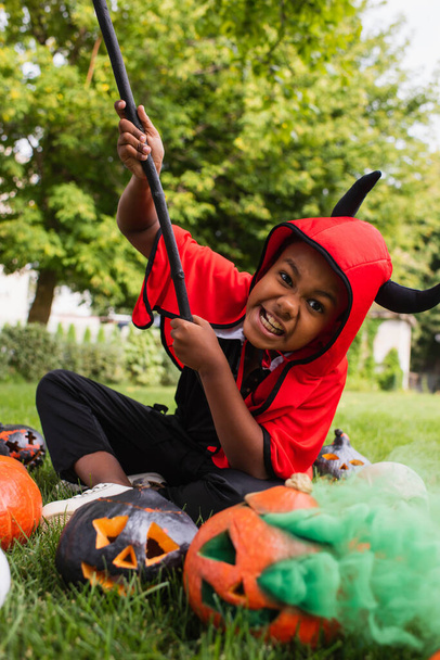 blurred african american boy in devil halloween costume grinning while holding broom near pumpkins and sitting on lawn  - Fotó, kép