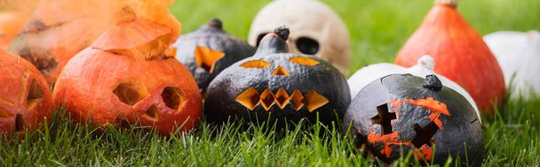 carved and scary pumpkins with orange smoke near blurred skull on green lawn, banner - Photo, Image