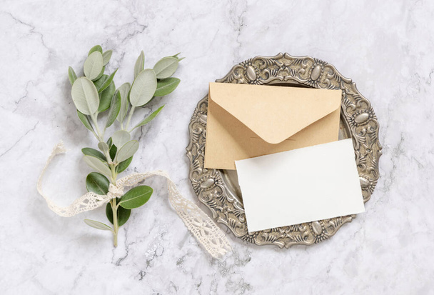 Порожня паперова картка та конверт з гілками евкаліпта на старовинній тарілці на мармуровому столі зверху. Макет картки
 - Фото, зображення