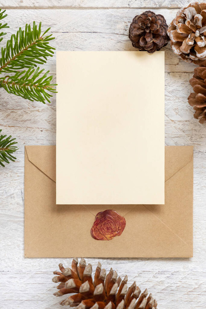 Winter Christmas Composition with a card over sealed envelope flat lay. Christmas and New Year greeting card template with fir tree branches and pine cones on white wooden background top view. Holiday mockup. - Photo, Image