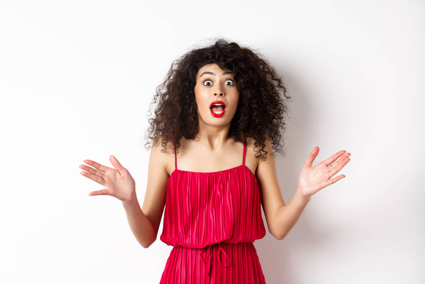 Excited woman with curly hair, talking with amazed face, explain big news, shaking hands and look astonished at camera, white background - 写真・画像