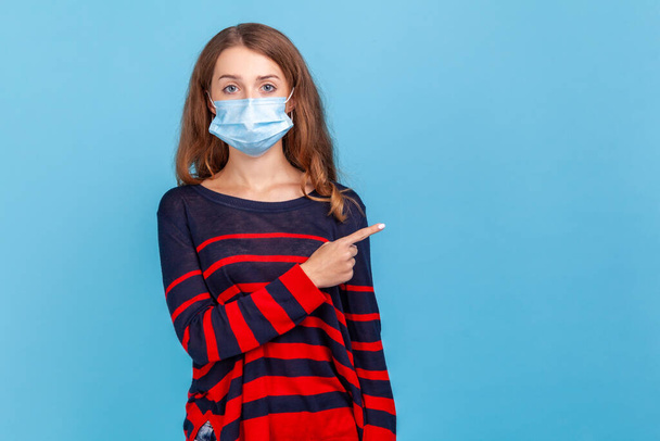 Portrait of serious woman wearing striped casual style sweater and surgical mask pointing finger side for empty wall for advertisement. Indoor studio shot isolated on blue background. - Photo, image