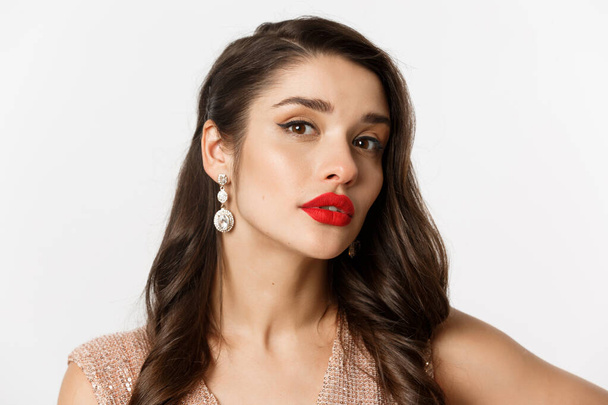 Concept of New Year celebration and winter holidays. Close-up of sensual and beautiful woman with red lips, looking confident at camera, wearing luxury earrings, white background - Photo, Image
