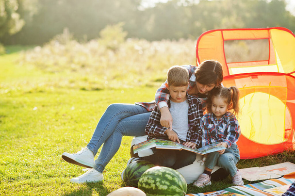 Family spending time together. Mother with two kids read book outdoor in picnic blanket and kids tent. - Photo, image