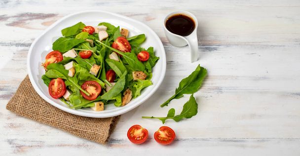 Verse spinazie salade met tomaat, kleine stukjes geroosterde kip in witte keramische schotel op bruine zak, samen met balsamico azijn op witte houten tafel. Concept voor gezond. - Foto, afbeelding