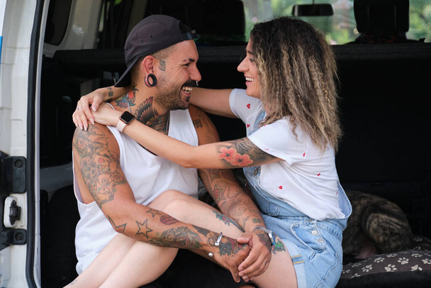 Young tattooed couple smile, hug and look each other in the back of the van. - Photo, Image