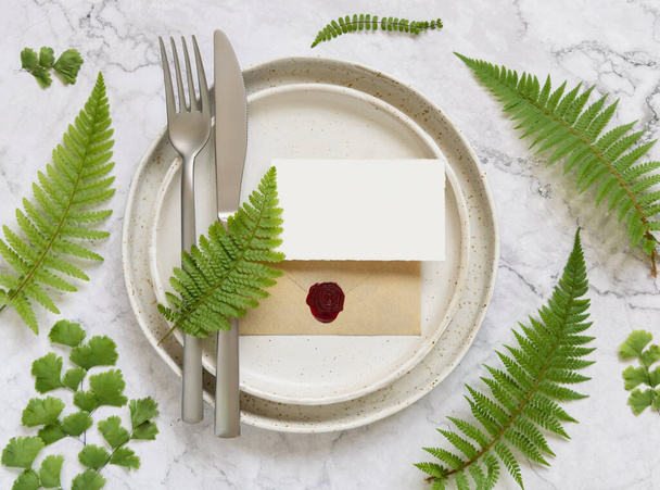 Blank paper card and sealed envelope on table setting decorated with fern leaves on white marble table top view. Tropical mock-up scene with place card flat lay - Photo, Image