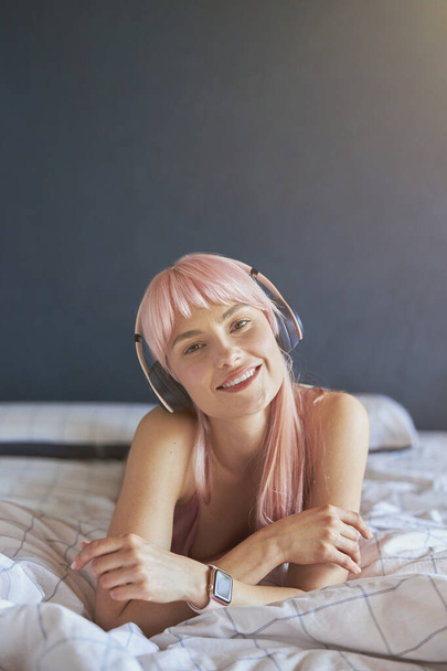 Портрет красивой модели с розовыми волосами, слушающей музыку на удобной кровати дома - Фото, изображение