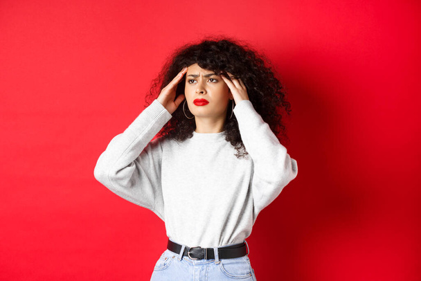 Distressed italian girl with curly hair, touching head temples and feeling bad, having headache, looking aside, standing on red background - Foto, Imagen