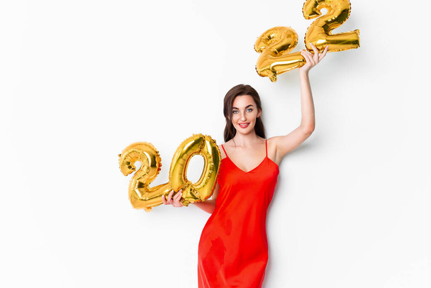 Young smiling woman in red cocktail dress with bright make-up celebrating New Year 2022 and holding golden balloons 2022 in hands on white background - Foto, afbeelding