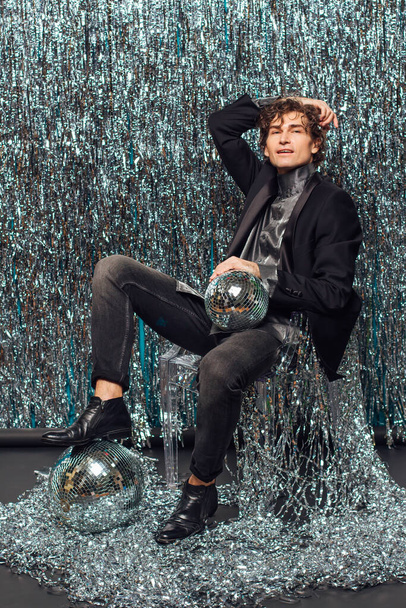 Tall attractive man sitting on the floor over the sparkle background and mirror disco ball holding glass of champagne. Party concept. - Valokuva, kuva