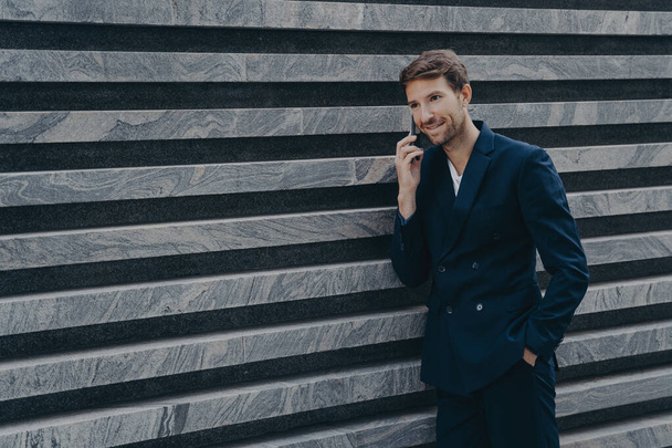 Trabajador corporativo masculino vestido con traje negro formal mantiene la mano en el bolsillo tiene conversación telefónica - Foto, Imagen