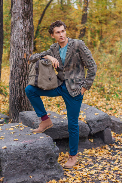 Tall handsome man dressed in a brown jacket posing with a bag near the rocks in autumn forest - Fotó, kép