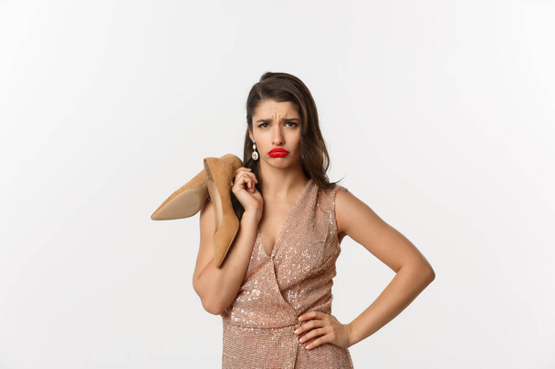 Party and celebration concept. Image of attractive clingy woman complaining, tired feet after wearing heels, holding shoes in hands and sulking, standing over white background - Fotografie, Obrázek