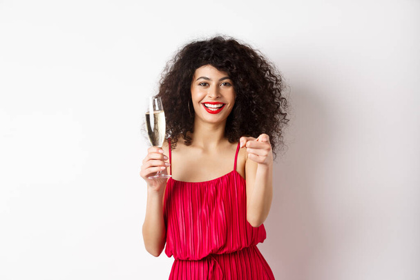Όμορφη σγουρή γυναίκα με κόκκινο φόρεμα, πάρτι ή έχοντας ρομαντική ημερομηνία, κρατώντας ένα ποτήρι σαμπάνια και δείχνοντας σας, καλώντας το πρόσωπο, στέκεται σε λευκό φόντο - Φωτογραφία, εικόνα