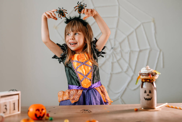 Portrait of little girl dressed Halloween witch costume and spider headband sitting at the table with sweets and monster shake or freak milkshake. Selective focus. - Foto, Imagem