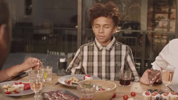 Skrzynia nastoletniego Afroamerykanina siedzącego przy stole, trzymającego się za ręce z przyciętymi członkami rodziny, modlącego się przed kolacją - Materiał filmowy, wideo