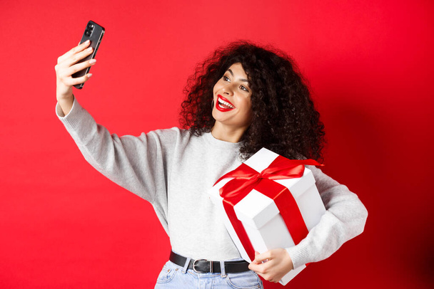 Διακοπές και τεχνολογική έννοια. Ευτυχισμένη γυναίκα που παίρνει selfie με το δώρο της, κρατώντας δώρο και smartphone, στέκεται σε κόκκινο φόντο - Φωτογραφία, εικόνα