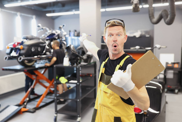 Verrast mannelijke reparateur met klembord met documenten en wijzende vinger naar motorfiets - Foto, afbeelding