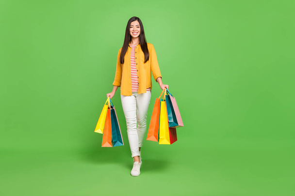 Full length photo of impressed brown hairdo young lady hold bags go wear shirt trousers sneakers isolated on green color background - Fotoğraf, Görsel