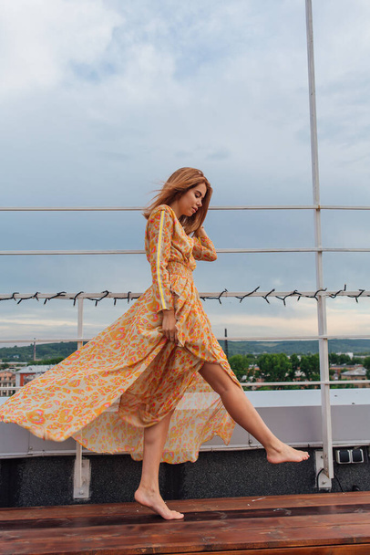 Beautiful young woman in long leopard dress standing with bare feet on the stairs on the top of the roof of a building. - Fotó, kép