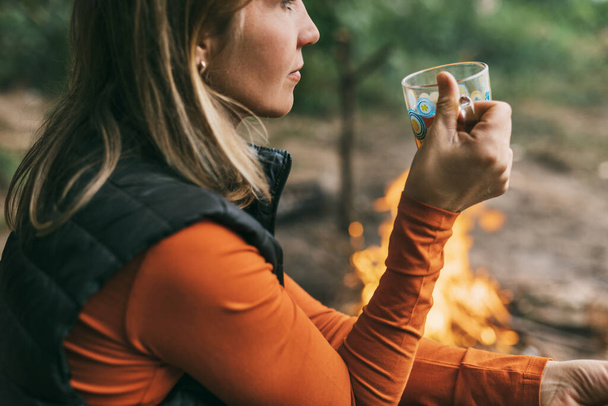 Közelkép egy felnőtt nőről, aki egy csésze teával üldögél a tűz mellett, miközben az erdőben pihen. - Fotó, kép
