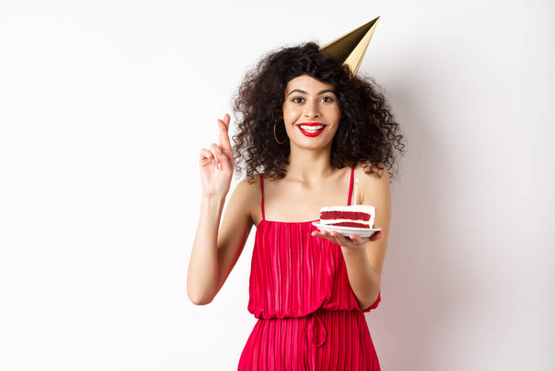 Возбужденная именинница празднует, загадывает желание, держит торт и скрещивает пальцы удачи, стоя счастливой на белом фоне - Фото, изображение