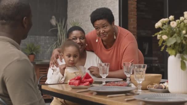 Inclinaison vers le bas de femme africaine mature étreignant sa fille adulte assise avec une jolie fille de cinq ans sur les genoux à la table du dîner, parlant à un homme méconnaissable - Séquence, vidéo
