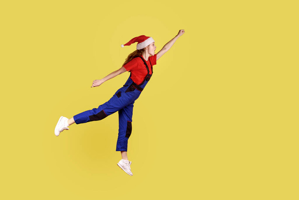 Full length profile portrait of handy woman flying like superhero, being hurry to serve all clients before Christmas, wearing overalls and santa hat. Indoor studio shot isolated on yellow background. - Фото, зображення