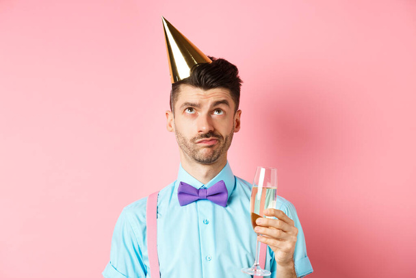 Holidays and celebration concept. Grumpy guy wearing birthday party hat and holding glass of champagne, looking up with skeptical face, standing on pink background - Photo, Image