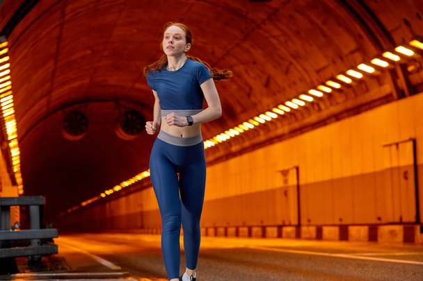 Fitness female runner in blue top and leggins confidently running along the road - Фото, изображение