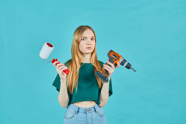 Beautiful girl with an electric screwdriver and an paint roller on blue background. Instruments accessories for renovation apartment room. Repair home concept. - Фото, изображение