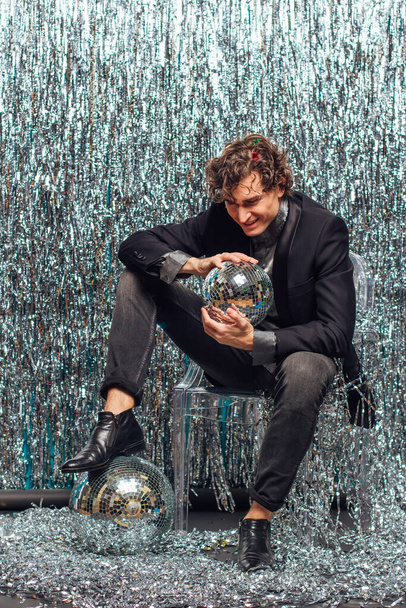 Tall attractive man sitting on the floor over the sparkle background and mirror disco ball holding glass of champagne. Party concept. - Foto, Imagen