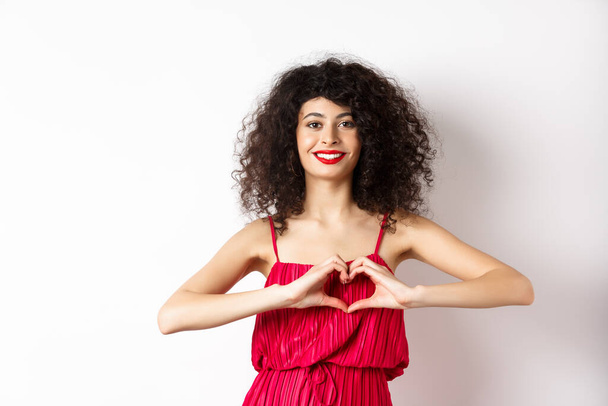 Encantadora joven con vestido romántico rojo, mostrando el gesto del corazón y sonriendo, expresar el amor, de pie sobre fondo blanco - Foto, imagen