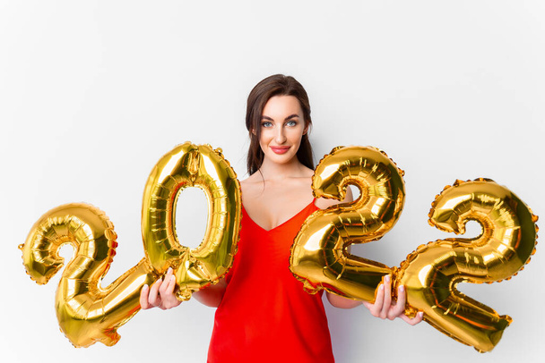 Young smiling woman in red cocktail dress with bright make-up celebrating New Year 2022 and holding golden balloons 2022 in hands on white background - Фото, зображення
