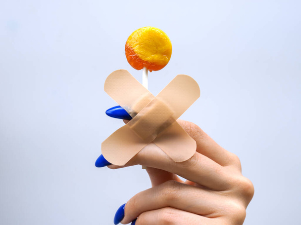 Γλυκός εθισμός. Γυναικείο χέρι με μπλε μανικιούρ που κρατά ένα κίτρινο γλειφιτζούρι. - Φωτογραφία, εικόνα