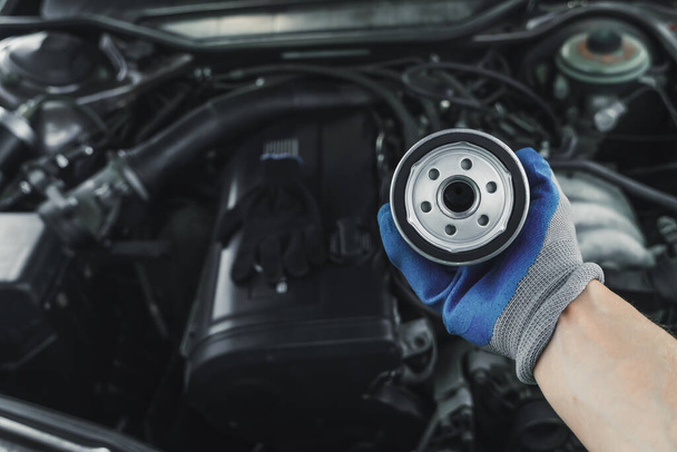 Sustitución del filtro de aceite. mecánico de mantenimiento del motor del coche sostiene un filtro de aceite en su mano contra el fondo de una campana de coche abierto - Foto, imagen